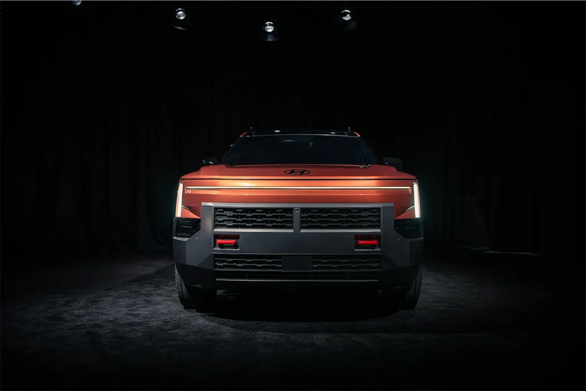 Front view of a striking orange vehicle with a modern, angular design, illuminated headlights, and a bold black grille, set against a dark background.