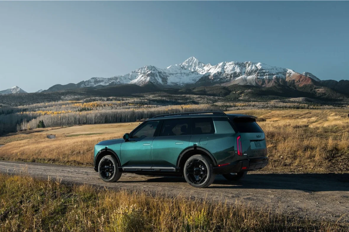 A teal SUV parked on a gravel road with a mountainous landscape in the background, featuring snow-capped peaks and autumn-colored trees.