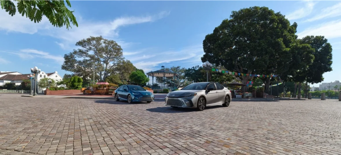 Two cars parked on a cobblestone square under a clear blue sky, with green trees and a decorative canopy in the background.
