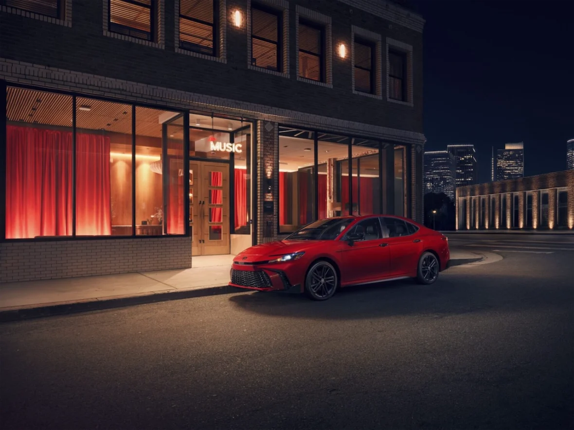 A bright red car parked on a street at night in front of a building with large windows, illuminated by warm light and featuring a sign that reads 'MUSIC'.