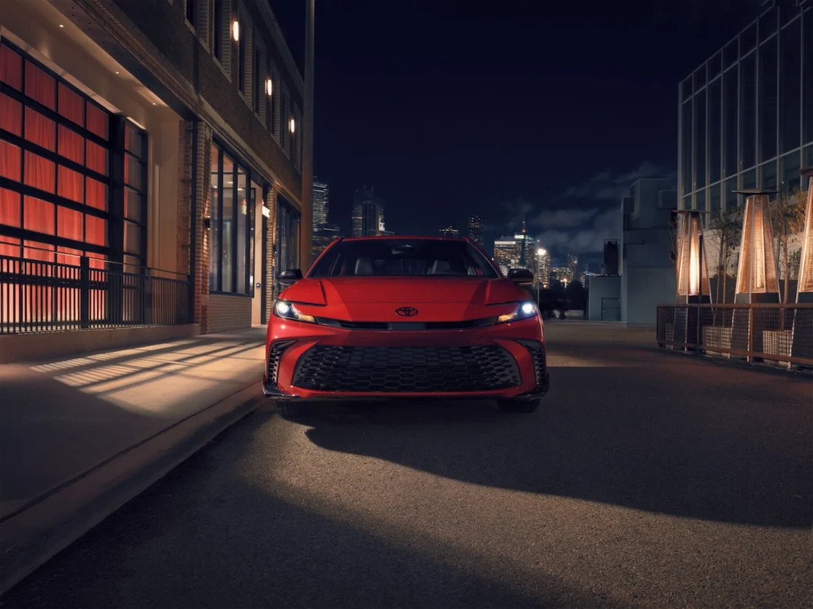 A red car parked on a city street at night, with modern buildings and city lights in the background.