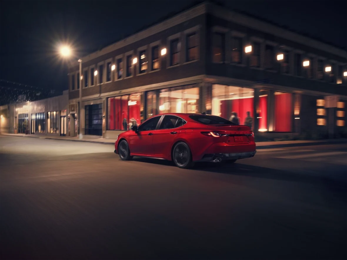A red car driving through a city street at night, illuminated by streetlights and nearby storefronts.