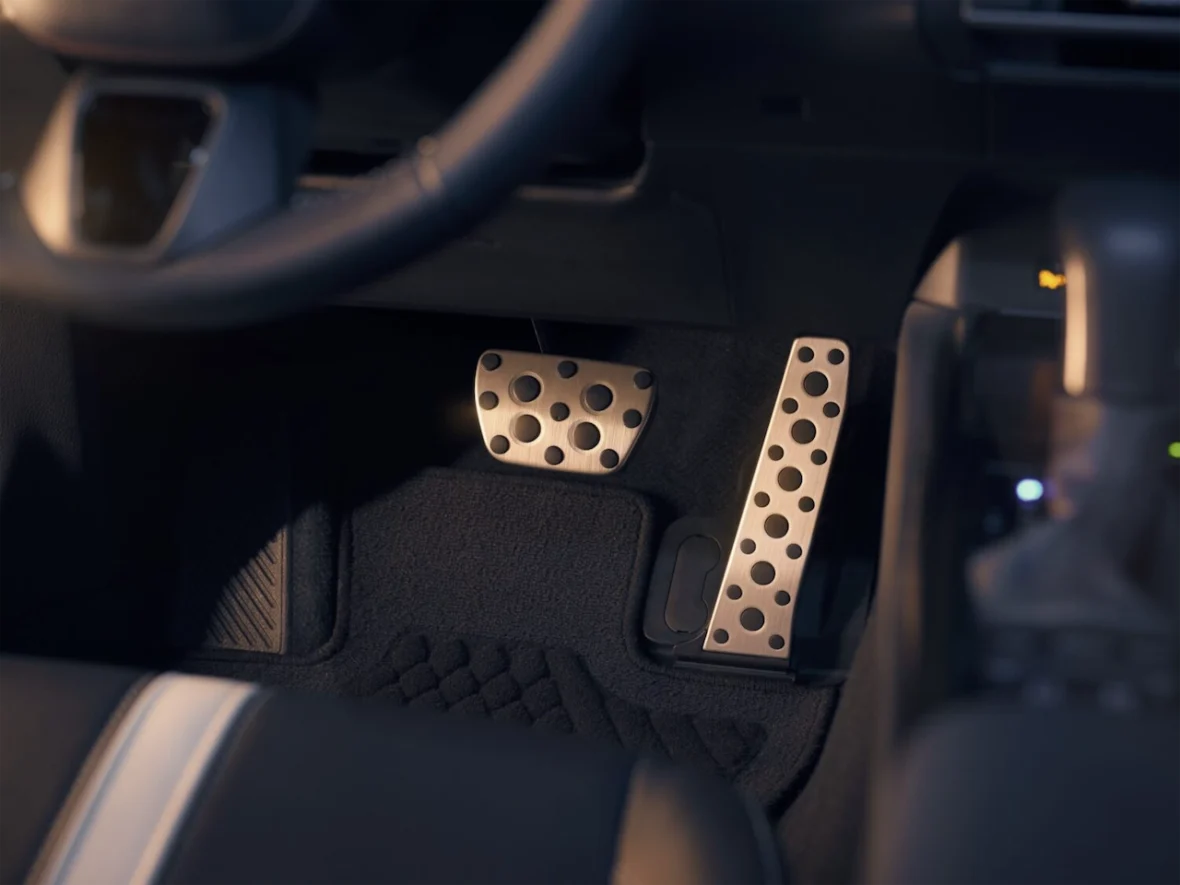 Close-up view of car floor pedals, including a metallic accelerator pedal with holes and a rubber brake pedal, shot from the driver's perspective.