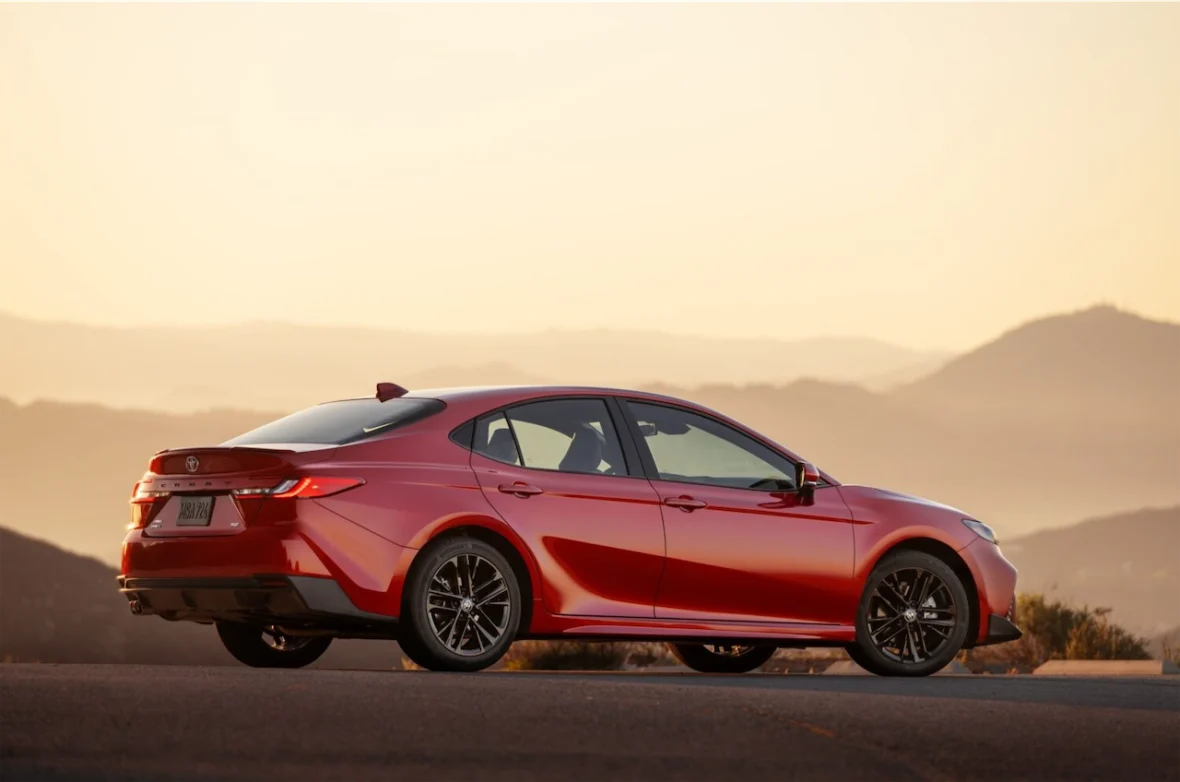 A red Toyota Camry parked on a scenic overlook, with mountains in the background and a warm sunset.