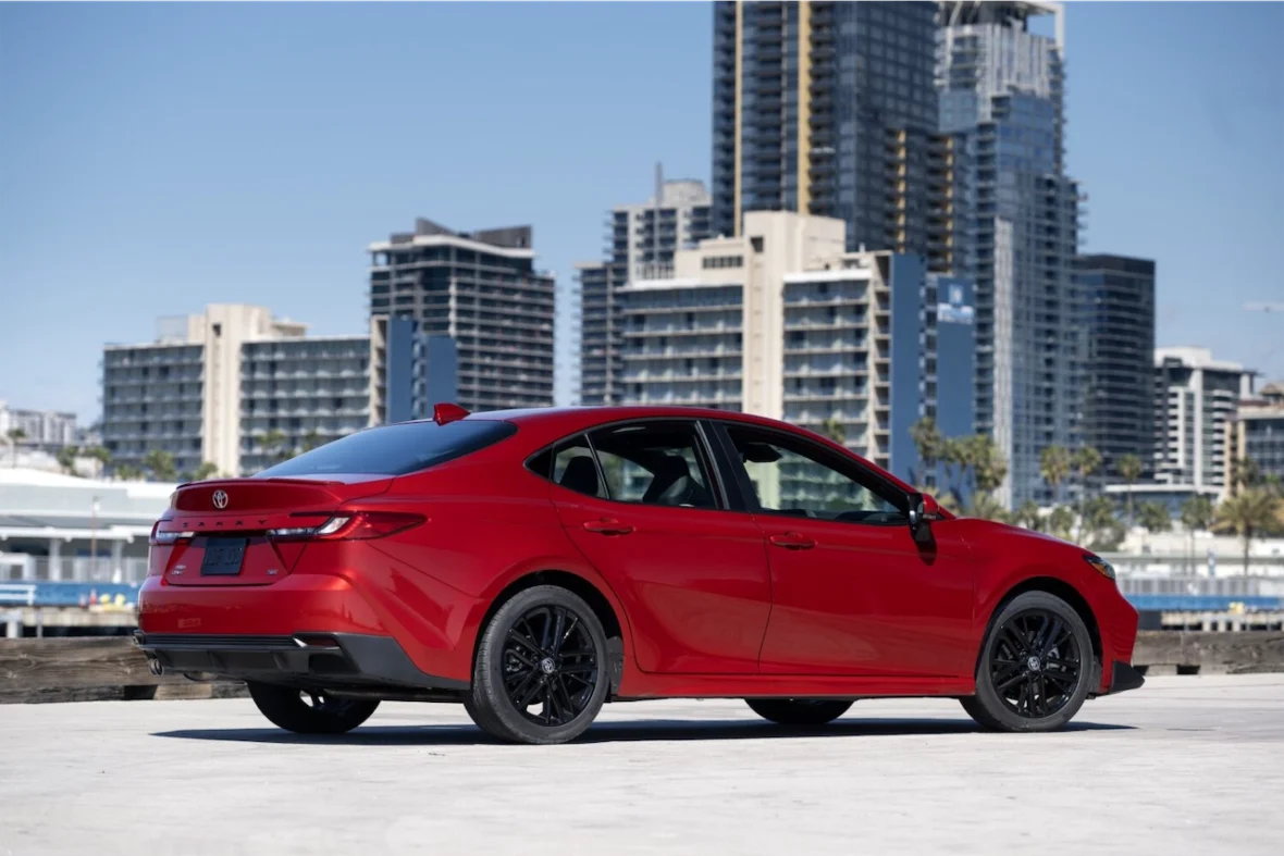 Red sedan parked by the waterfront with high-rise buildings in the background.