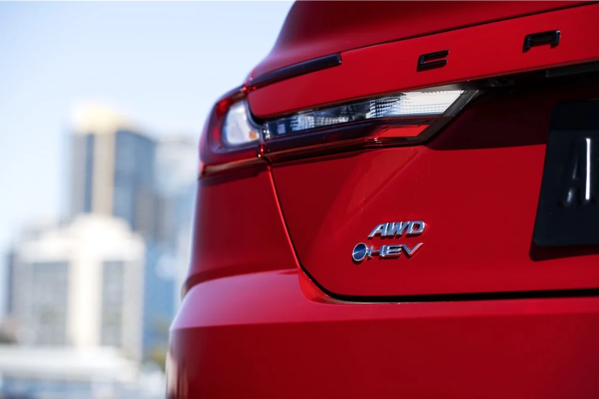 Close-up of the rear of a red car featuring the 'AWD HEV' badge with city buildings blurred in the background.