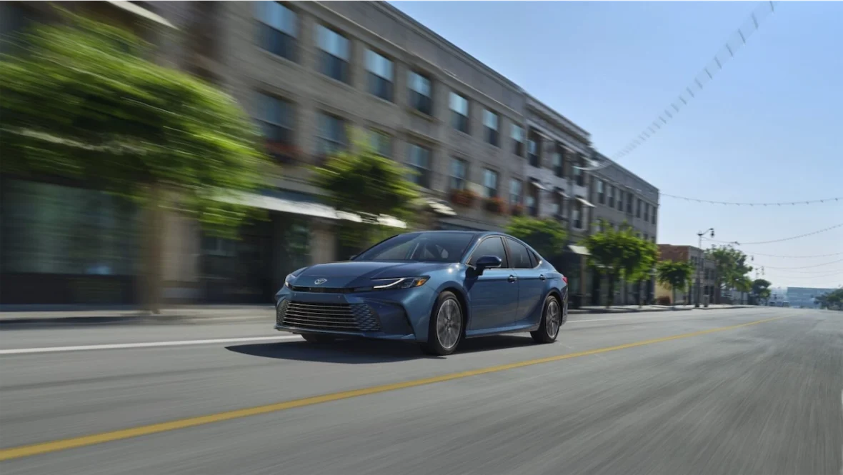 A blue sedan driving swiftly on a quiet city street with blurred trees and buildings in the background.