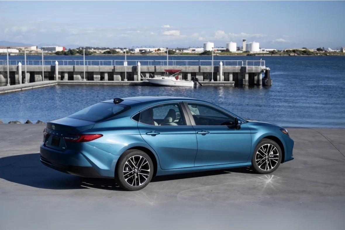 A blue sedan parked near a waterfront, with a marina and distant industrial buildings in the background.