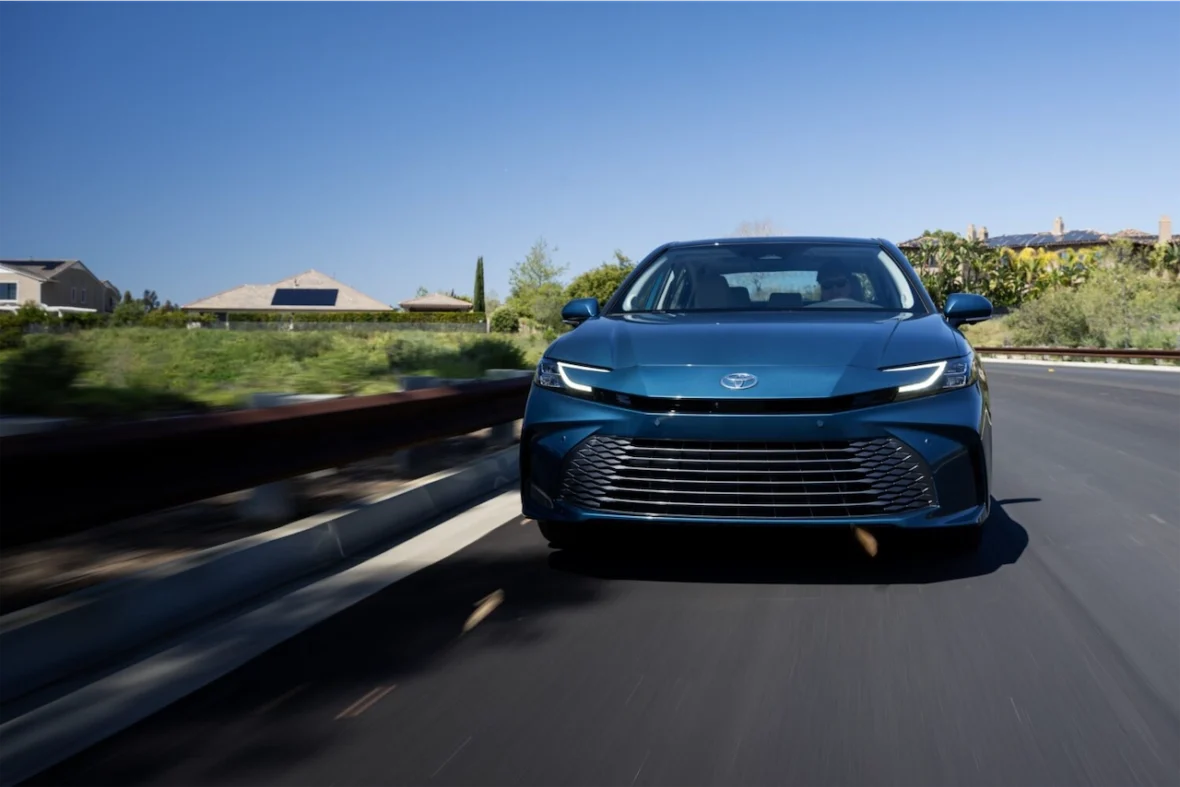 A blue Toyota sedan driving on a winding road with houses and greenery in the background under a clear blue sky.