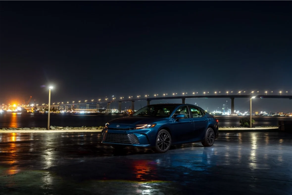 A blue sedan parked near a reflecting waterfront at night, with city lights and a bridge illuminated in the background.