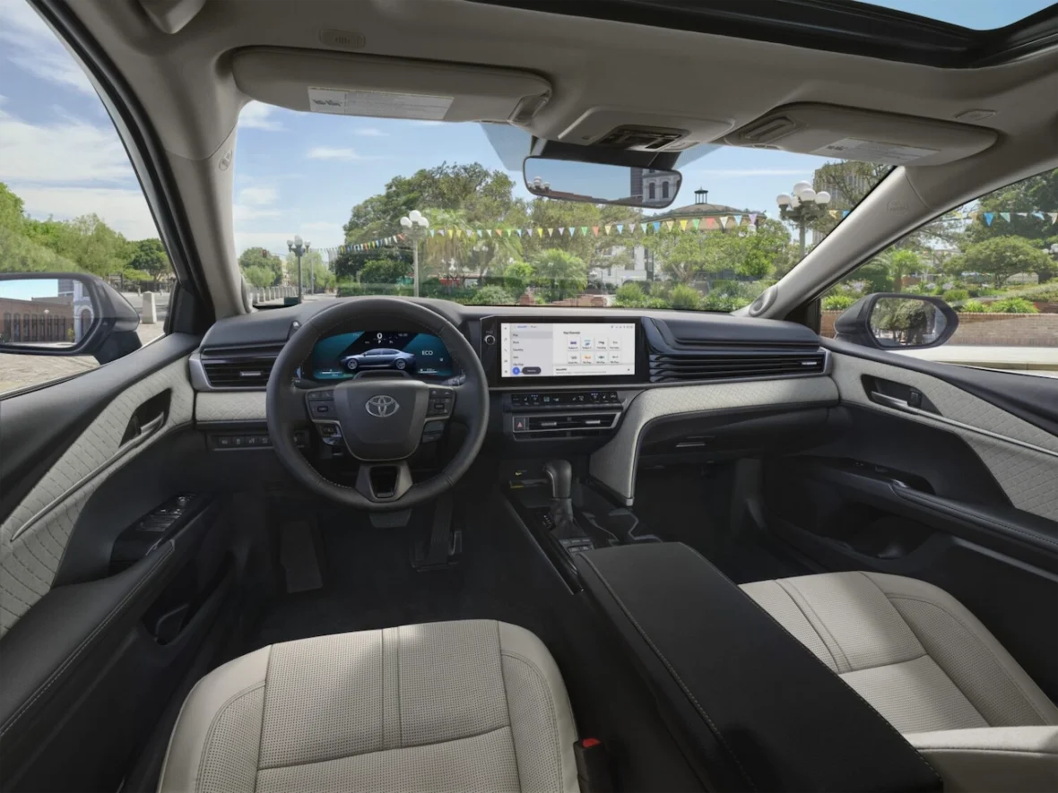 Interior view of a modern car with a Toyota dashboard, featuring a steering wheel, touchscreen display, and light-colored upholstery. A park with greenery is visible outside the windshield.
