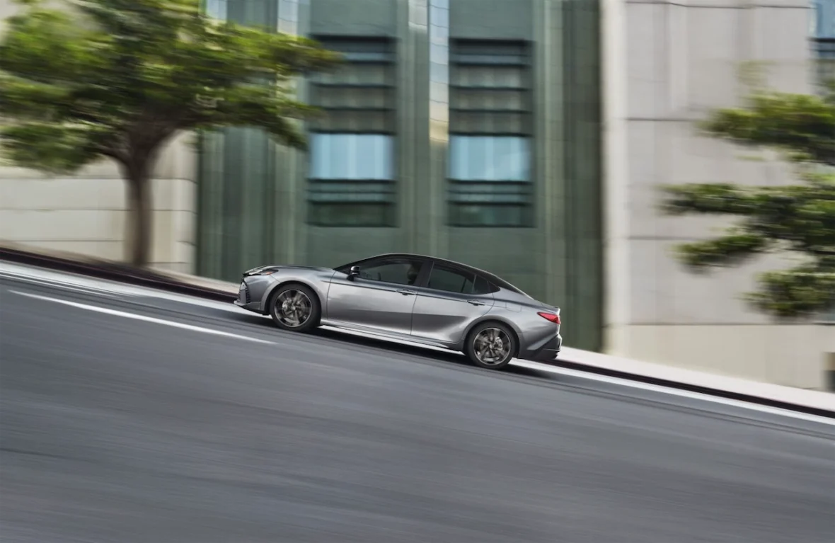 A silver sedan driving down a steep street with blurred trees and buildings in the background.