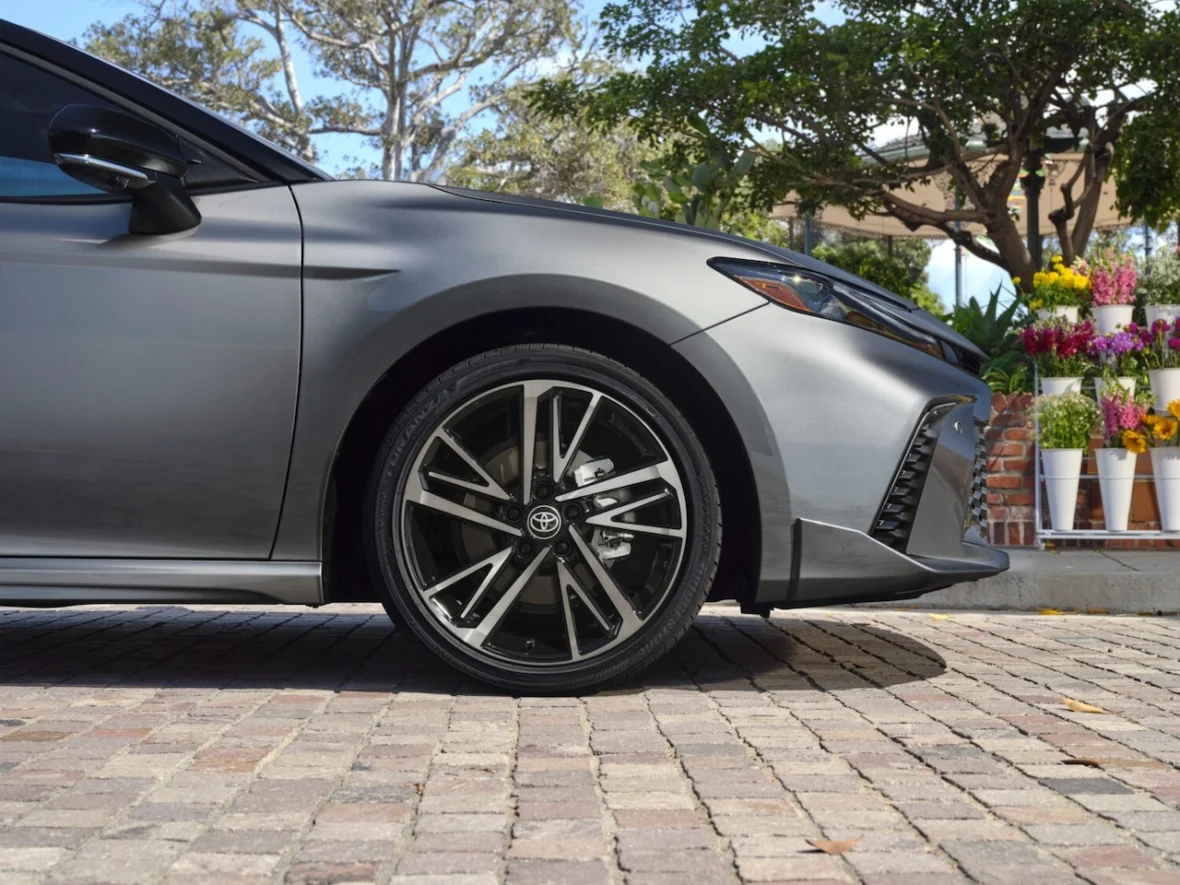 Close-up view of the side of a gray car with stylish alloy wheels, parked on a brick pavement. In the background, there are plants and flowers in pots.