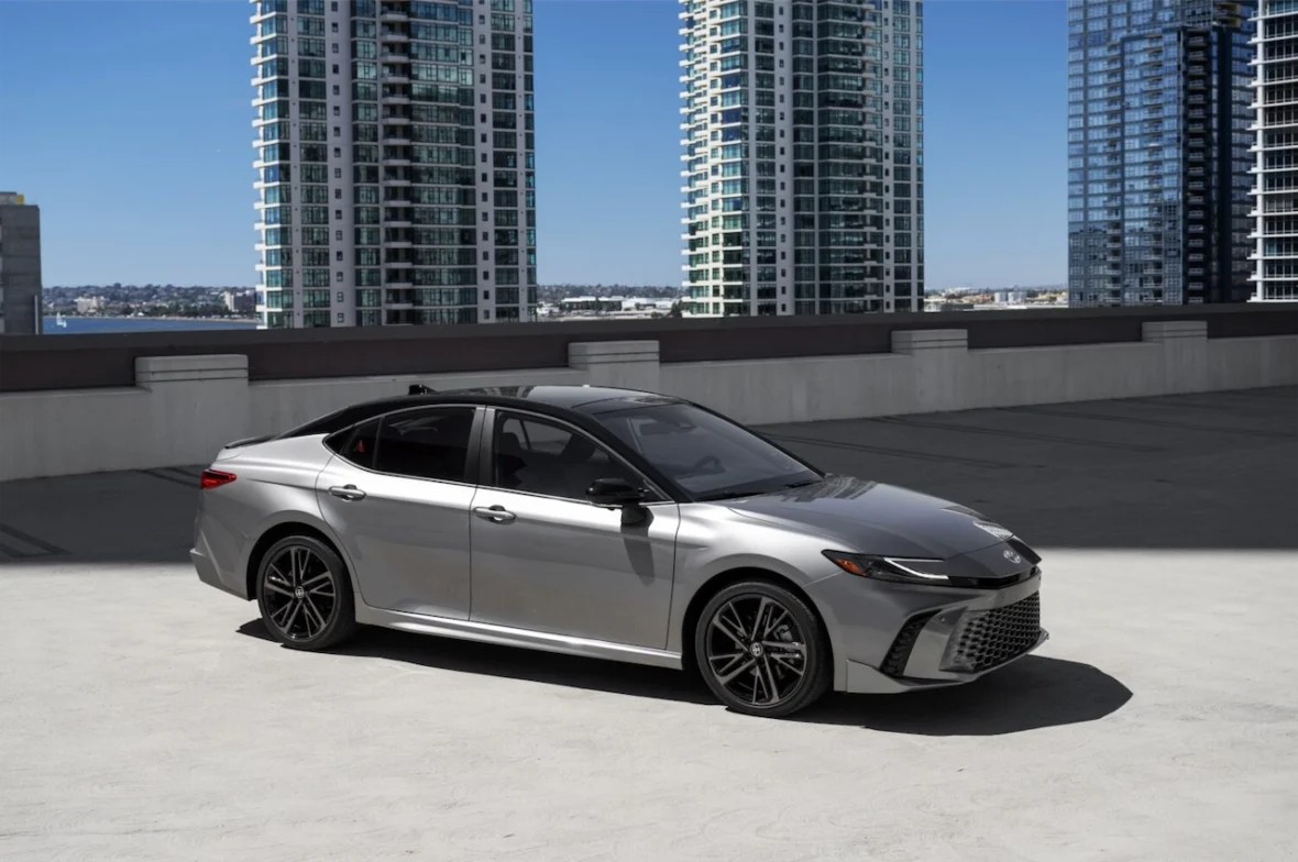 A silver sedan parked on a rooftop with modern high-rise buildings in the background under a clear blue sky.