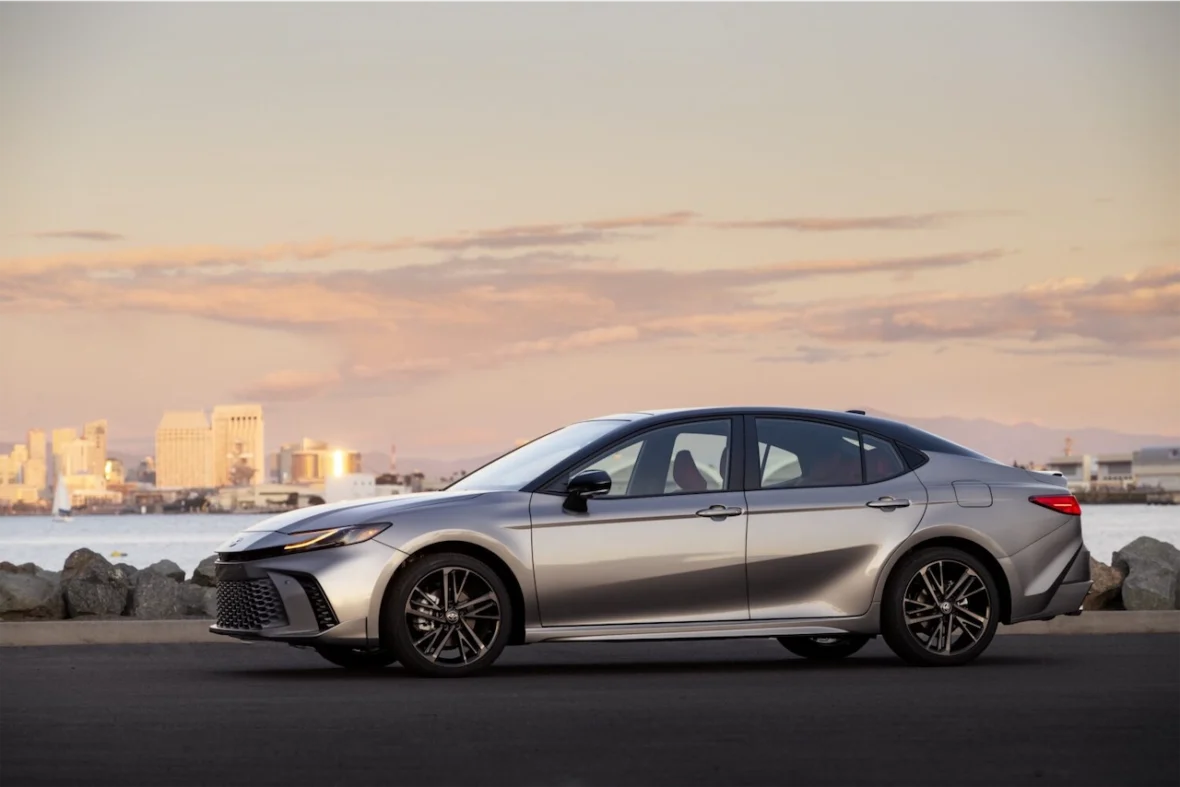 A silver sedan parked along a waterfront with a city skyline in the background at sunset.
