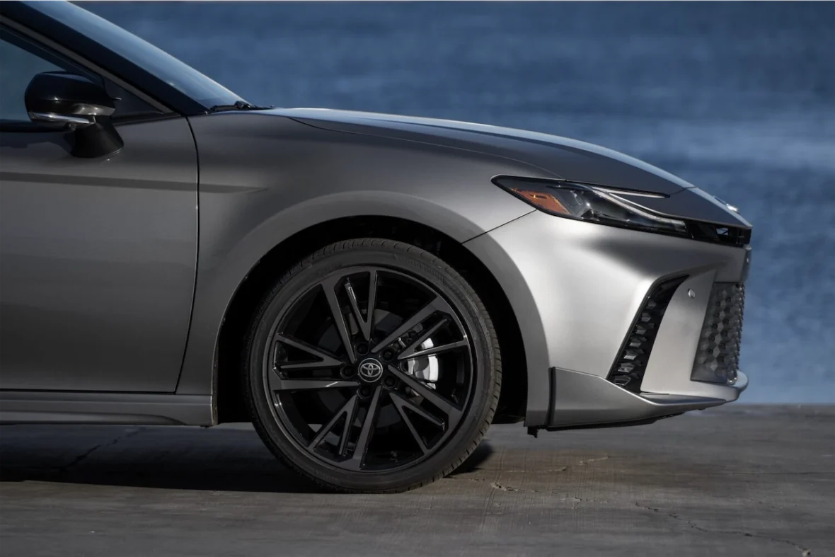Close-up of the front side of a silver car with a sleek design and black alloy wheels, set against a blurred background of water.