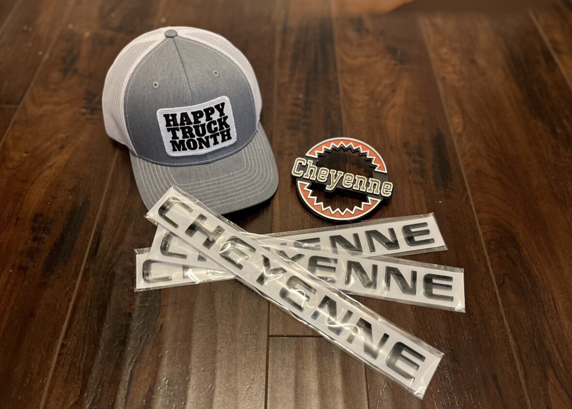 A gray and white cap with 'HAPPY TRUCK MONTH' printed on the front, accompanied by a round Cheyenne logo patch and several 'CHEYENNE' nameplates in plastic packaging, all displayed on a wooden surface.