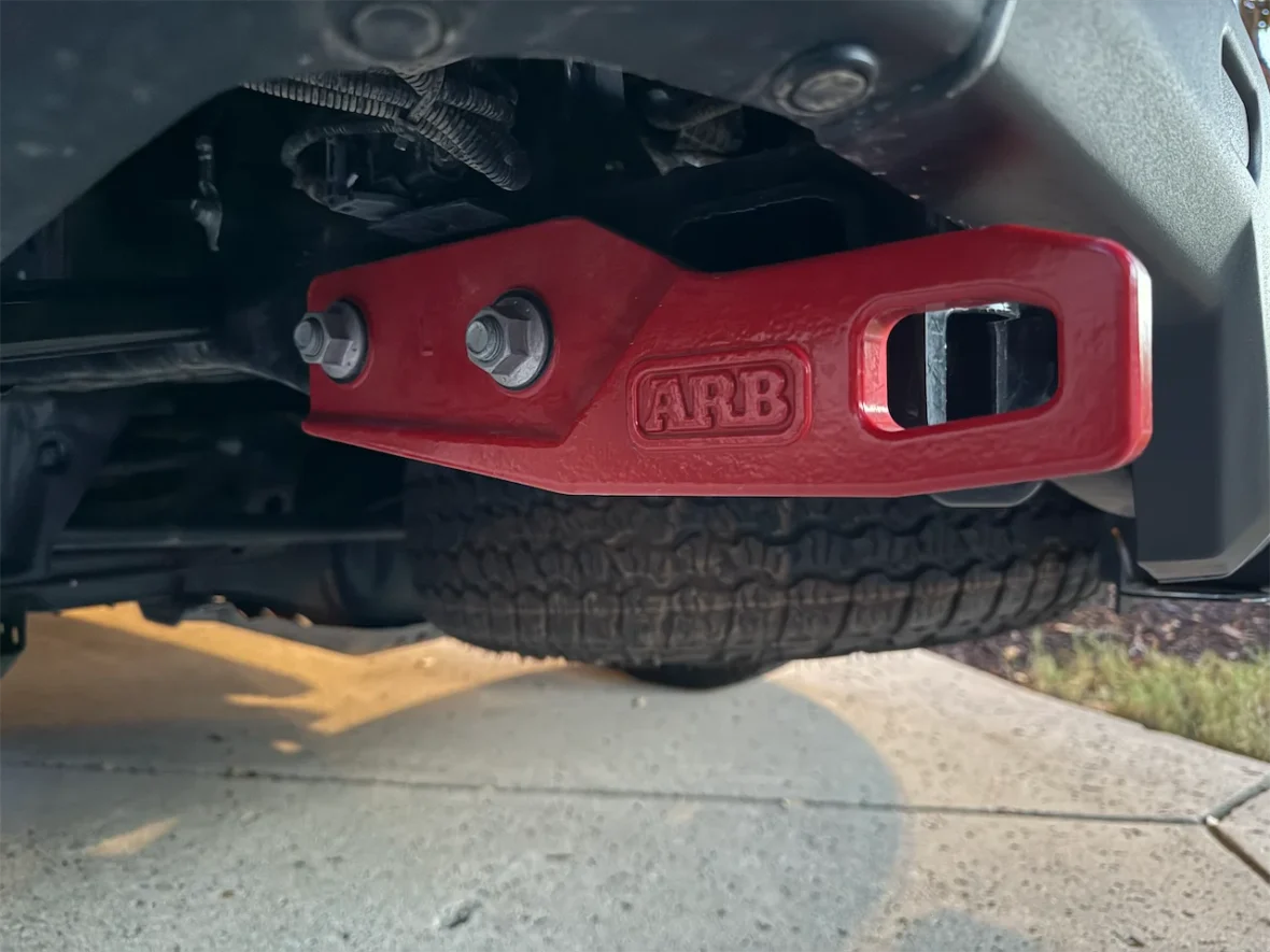 Close-up view of a red ARB bumper attachment with visible bolts, mounted under a vehicle next to a tire.