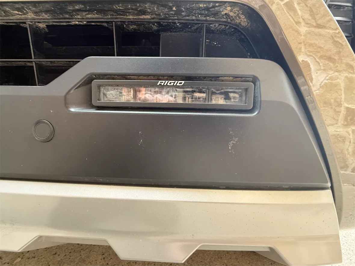 Close-up of a black vehicle bumper featuring a Rigid brand LED light bar and a round sensor.