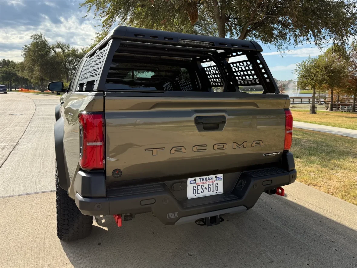 Rear view of a Toyota Tacoma pickup truck with a specialized bed cover, parked on a paved road surrounded by grass and trees.