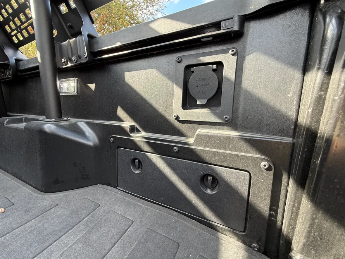 Interior view of a vehicle showing a textured black panel with a power outlet and a storage compartment.
