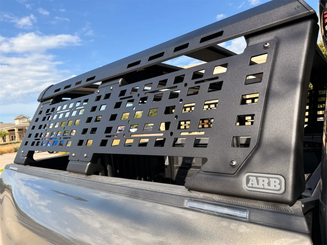 Close-up of a black ARB truck accessories rack installed in the bed of a pickup truck, showcasing its perforated design and a clear sky in the background.