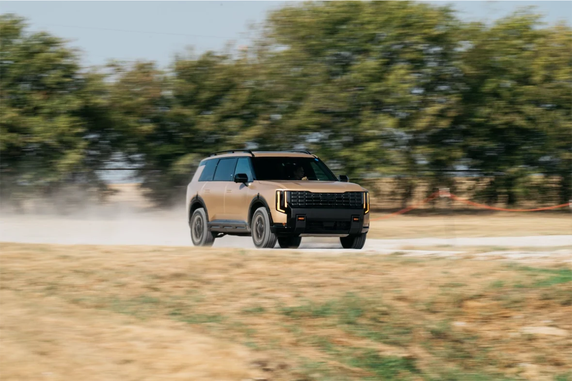 A tan SUV driving swiftly on a dirt road, creating a cloud of dust in its wake, with green trees in the background.
