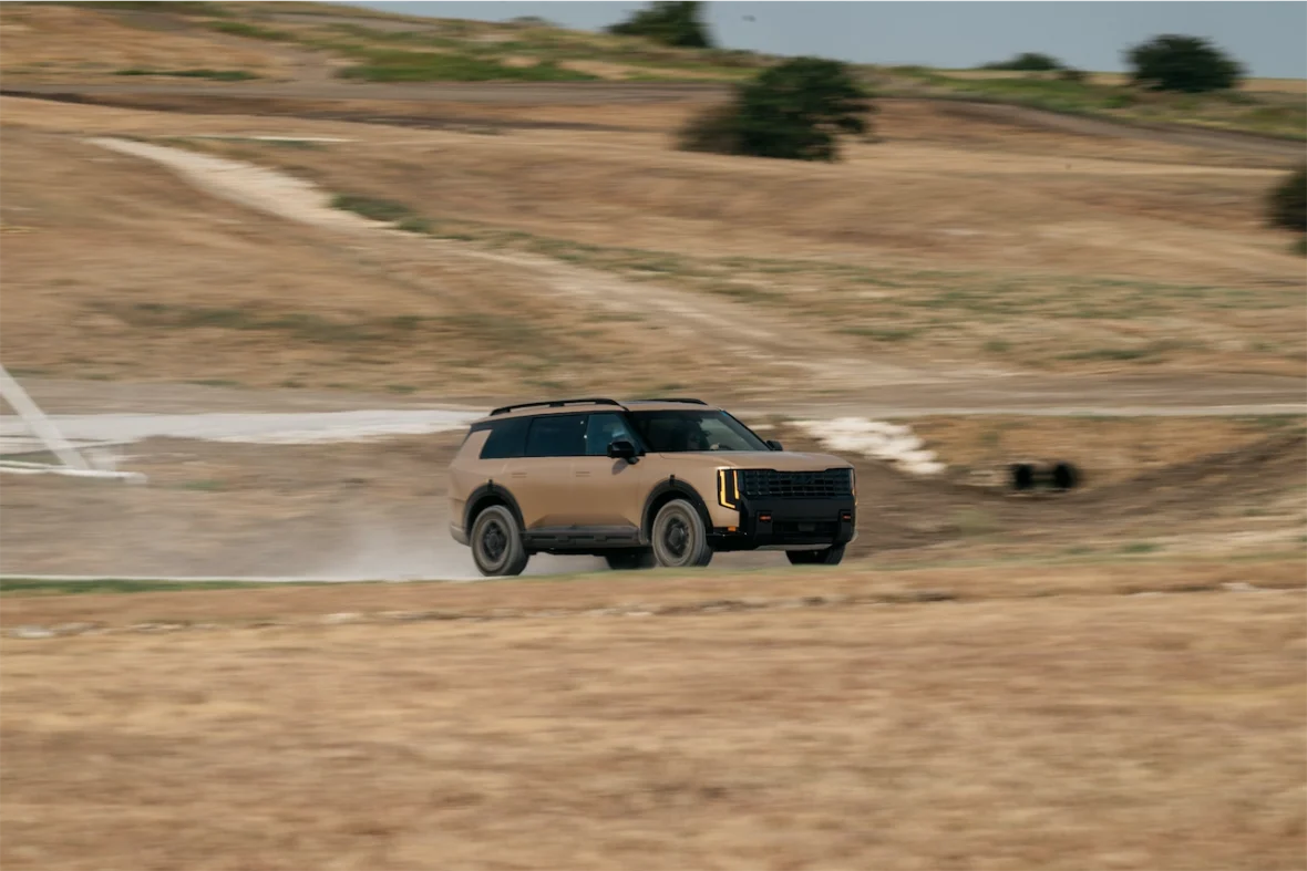 A modern SUV speeding across a dirt track in a rural landscape, with dry grass and hills in the background.