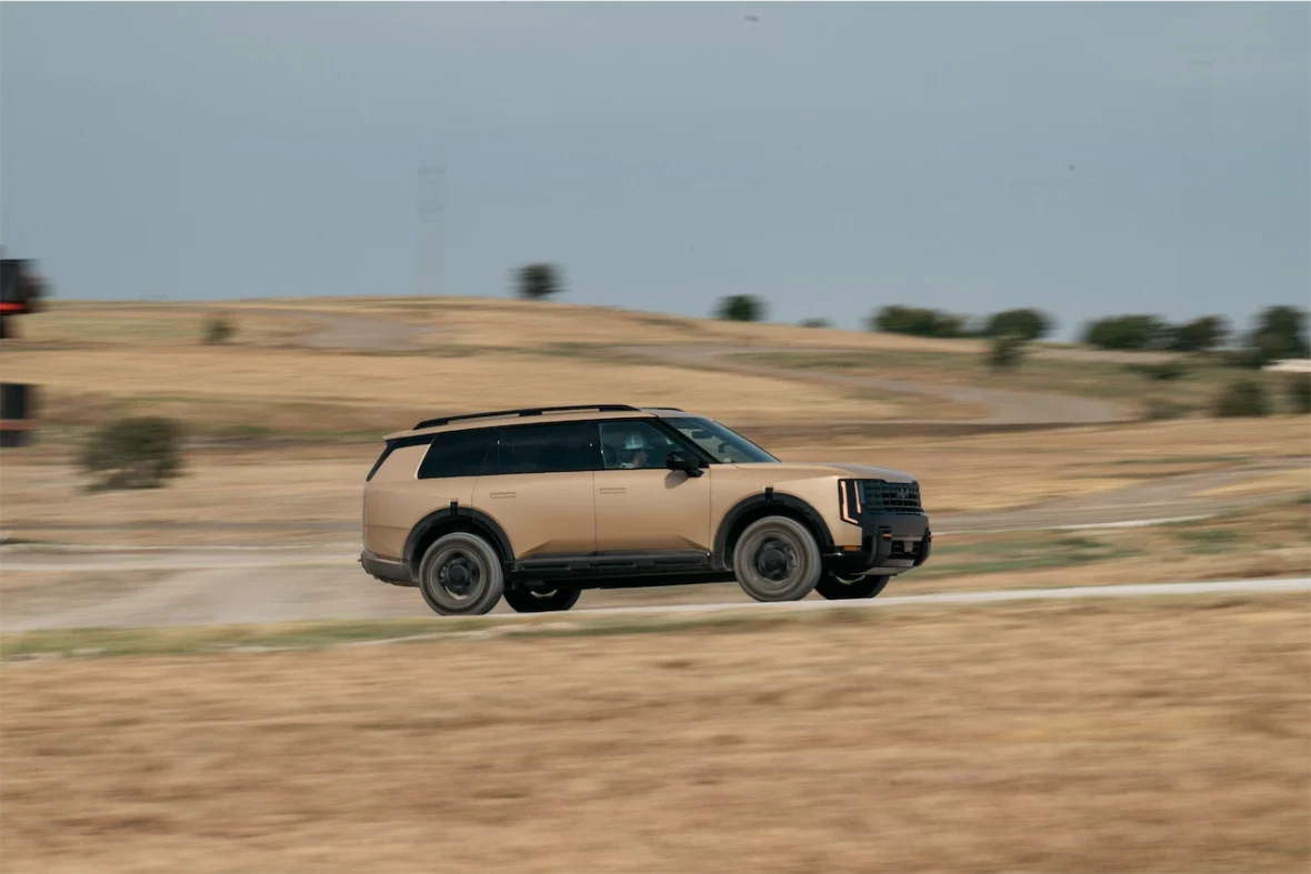 A beige SUV speeding along a dirt road in a rural landscape with dry grass and distant hills.