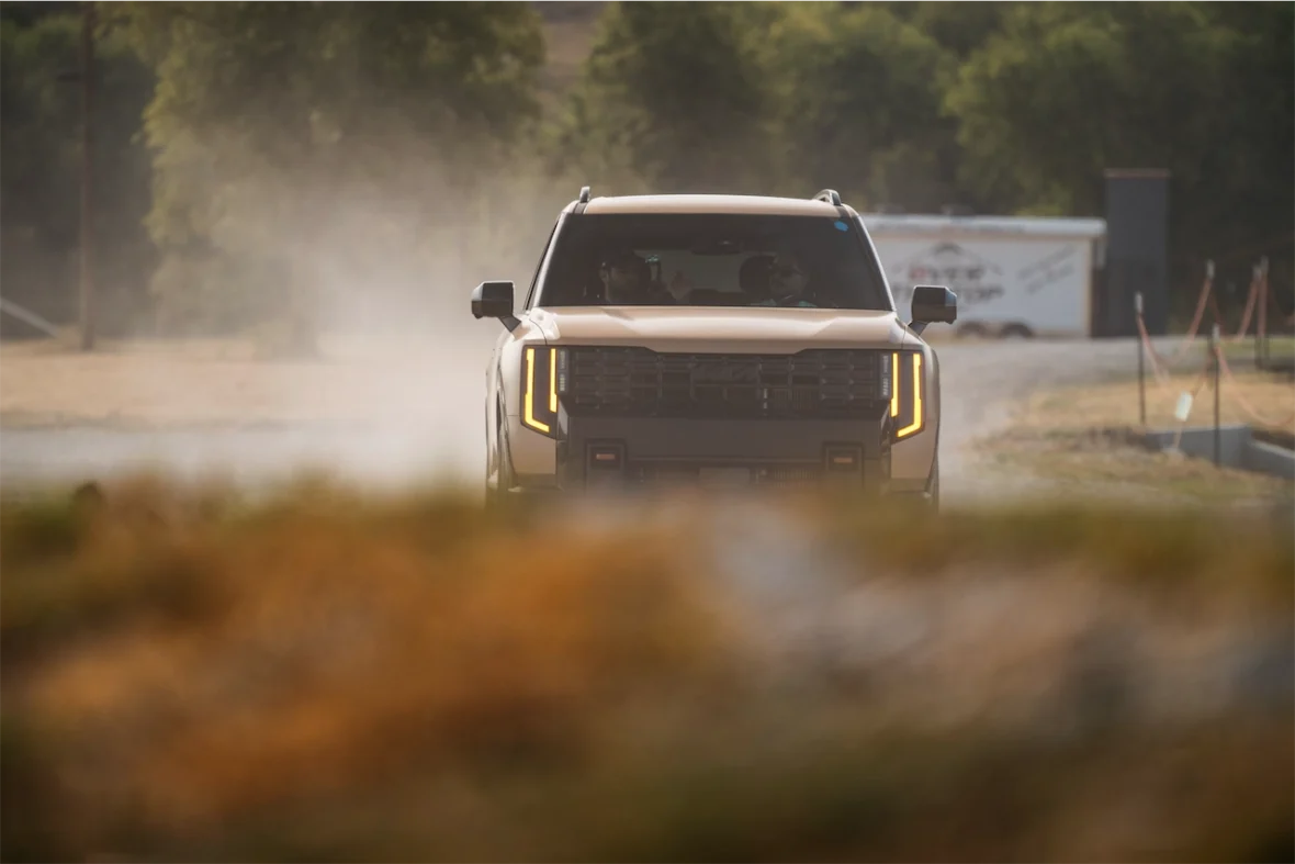 A front view of a tan SUV driving through a dusty road with trees in the background.