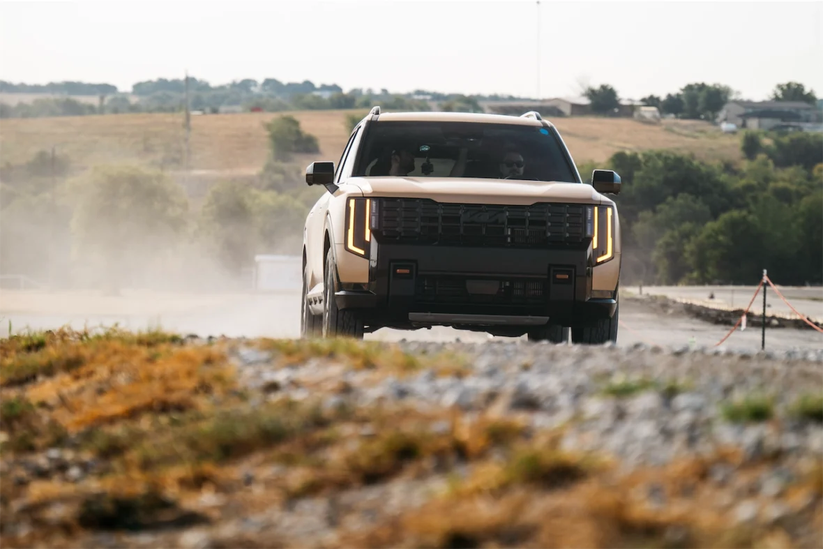 A modern SUV driving on a dirt road, kicking up dust in the background, with a scenic landscape of hills and trees.
