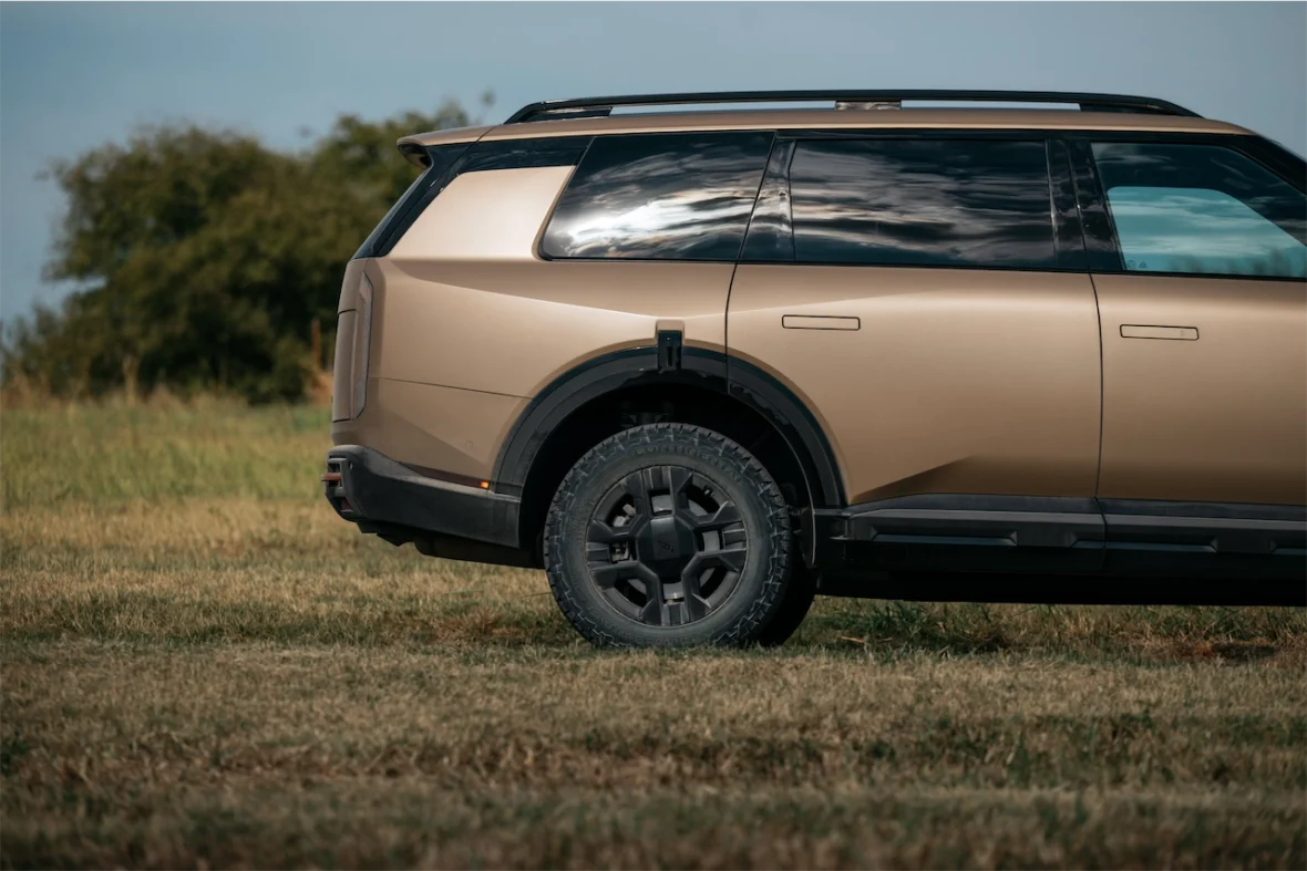 Side view of a modern SUV in a metallic bronze color parked on grassy terrain. The vehicle features a rugged tire and sleek design elements, with a clear blue sky in the background.
