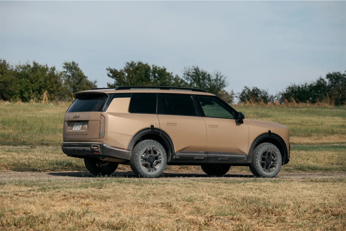 A beige SUV parked on a gravel surface, showcasing rugged tires and a modern design, set against a grassy field with trees in the background.