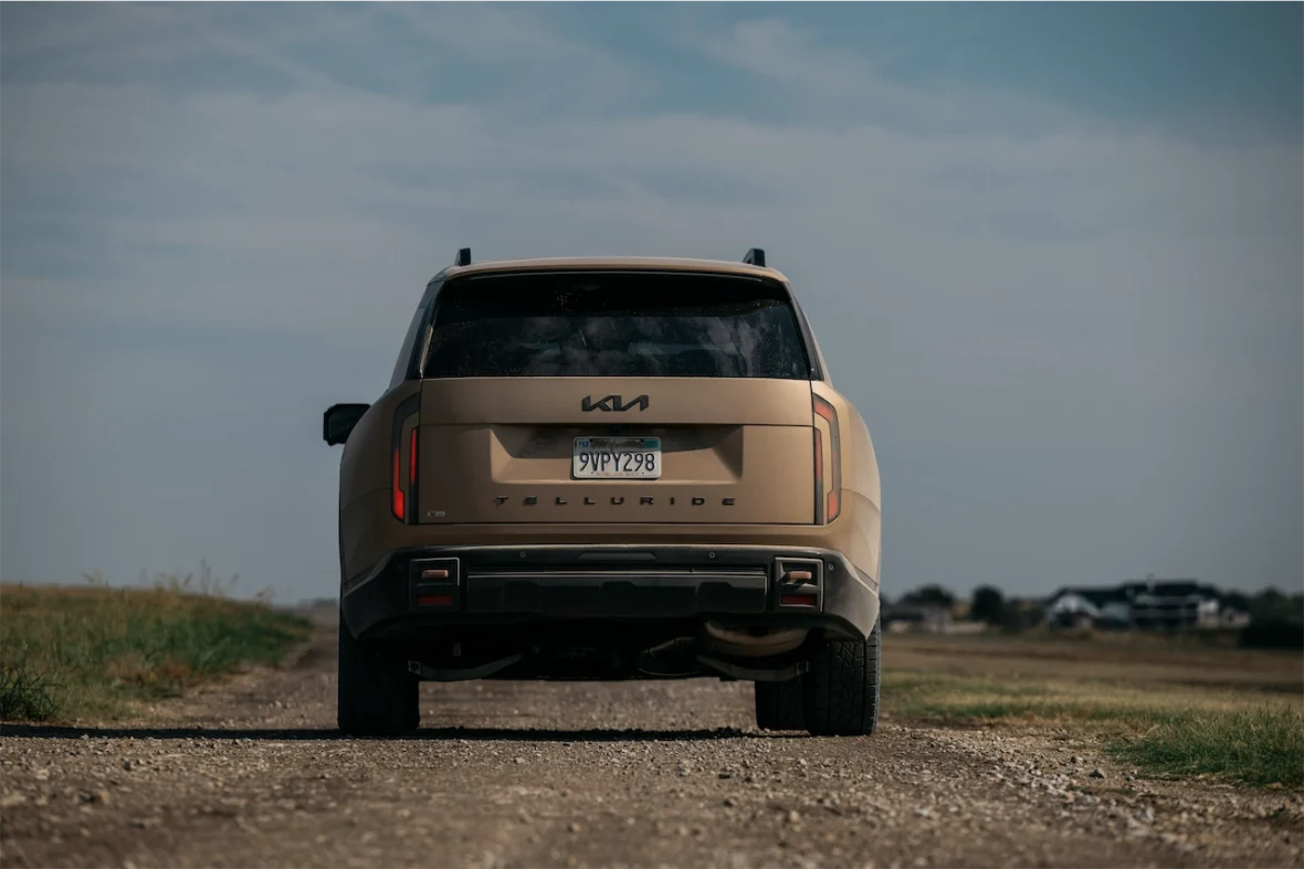 Rear view of a Kia Telluride parked on a gravel road with open fields and a cloudy sky in the background.