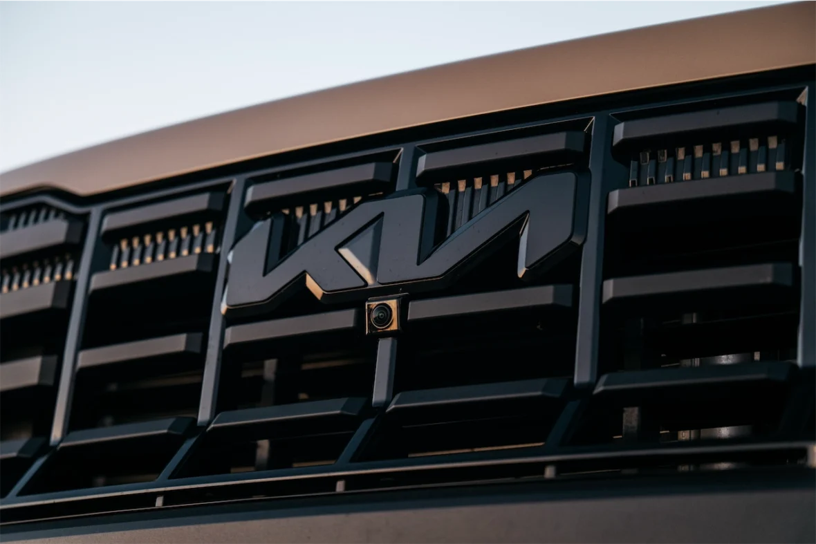 Close-up of a black Kia logo on a vehicle's grille, featuring a camera embedded in the design.