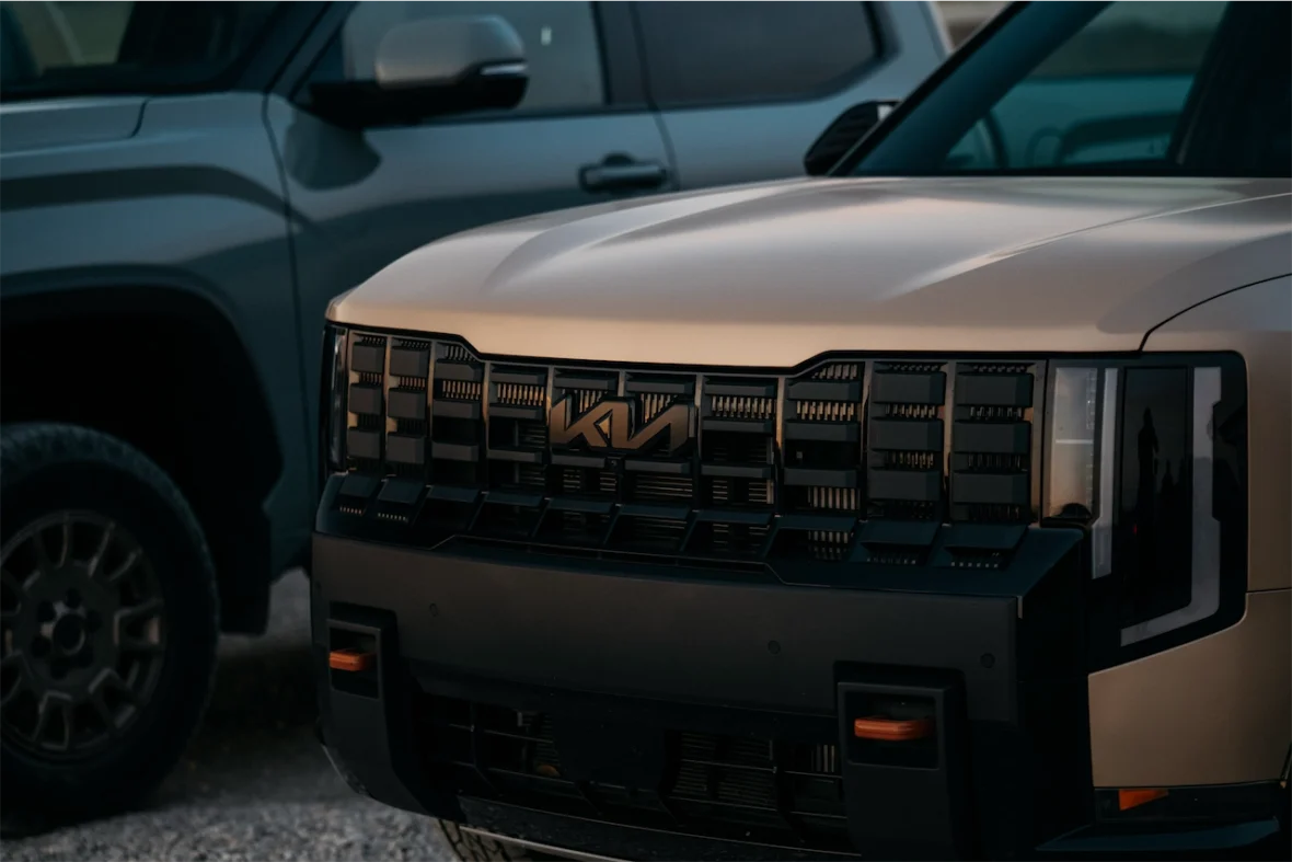 Close-up view of the front grille of a Kia vehicle, featuring a bold design and the Kia logo, with another vehicle partially visible in the background.