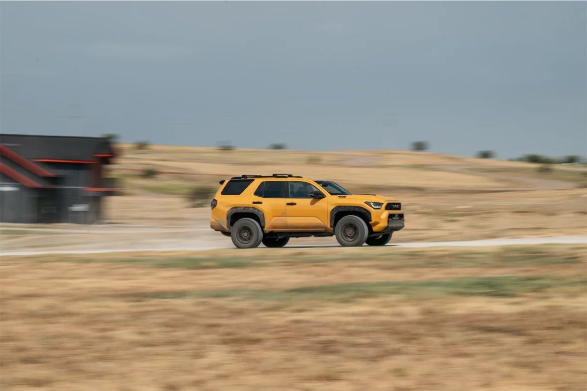 An orange SUV driving on a dirt road in a rural landscape with a house in the background.