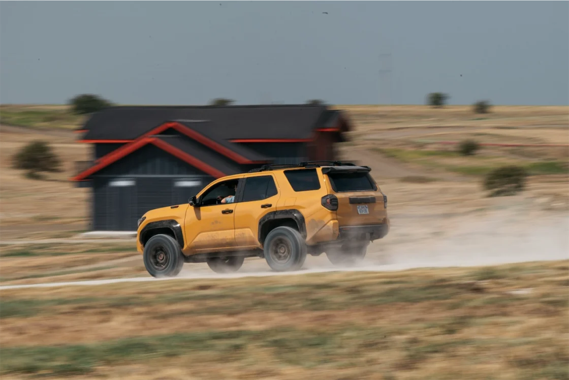 A yellow SUV is driving off-road on a dirt path, kicking up dust, with a modern black house in the background.