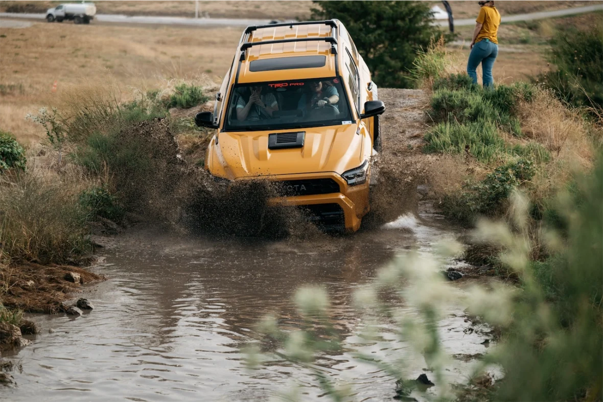 A bright yellow off-road vehicle splashing through a muddy puddle in a natural environment, with tall grass and a person observing in the background.