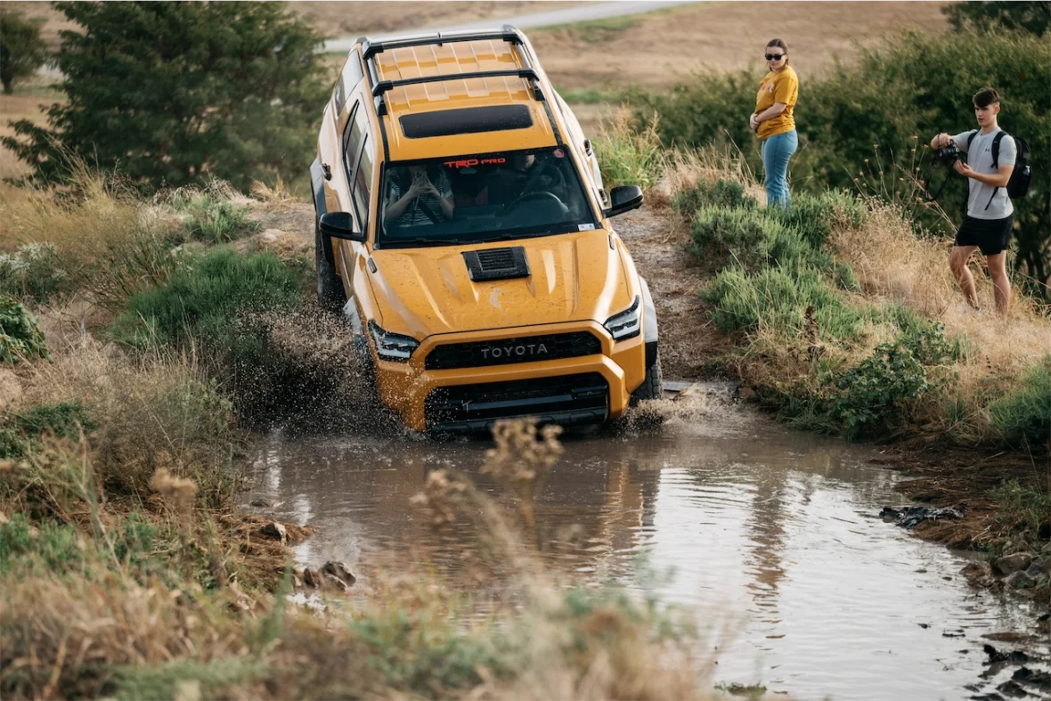 A yellow Toyota vehicle driving through a shallow water puddle on a gravel path, with a woman standing nearby observing and a man taking photographs.