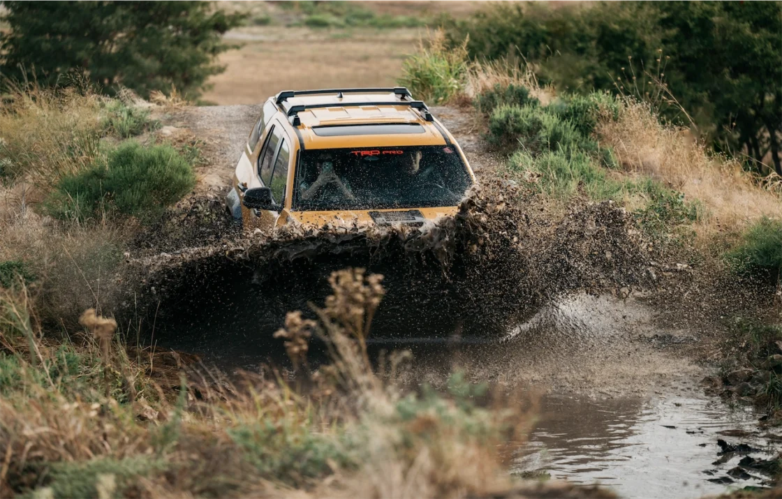 A yellow SUV splashing through a muddy water crossing surrounded by brush.
