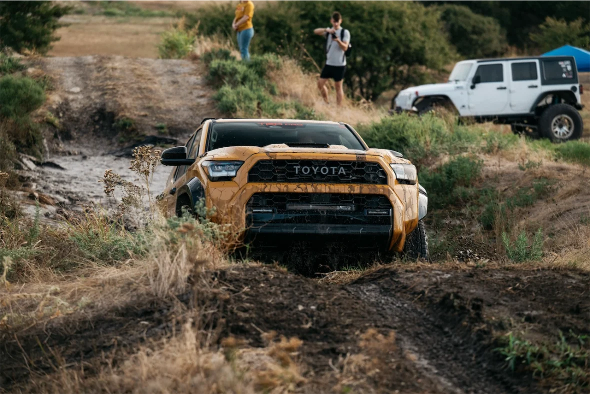 A yellow Toyota truck navigating through uneven terrain, with muddy patches and sparse vegetation. In the background, a white jeep and two people are visible.