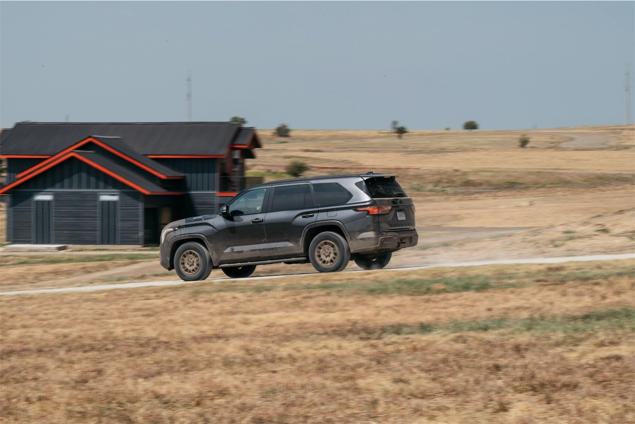 Gray SUV driving on a gravel road past a dark building
