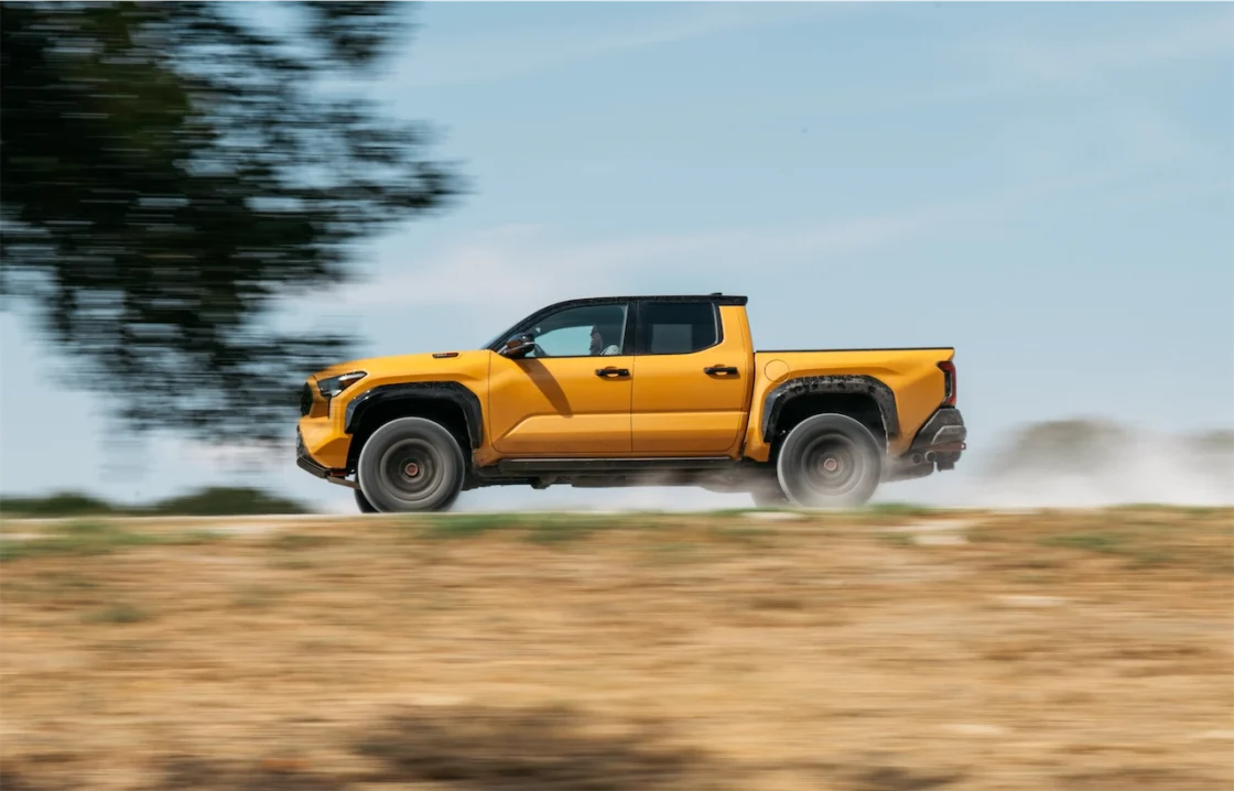 A bright yellow pickup truck driving swiftly on a dirt road, kicking up dust in the background.