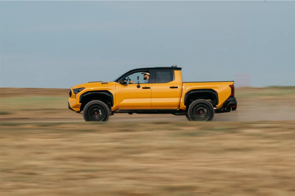 A bright yellow pickup truck speeding across a dirt field with a driver visible in the cabin.
