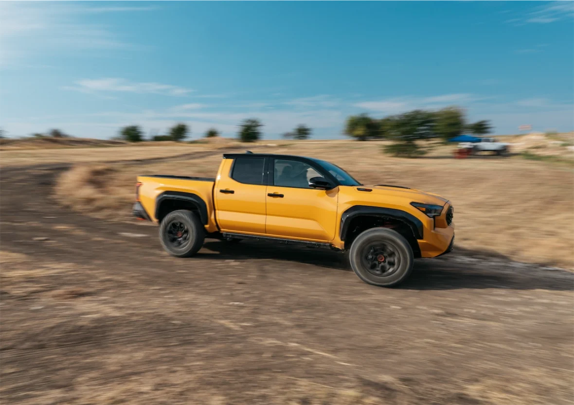 A yellow pickup truck driving on a dirt road surrounded by dry grass and trees under a clear blue sky.