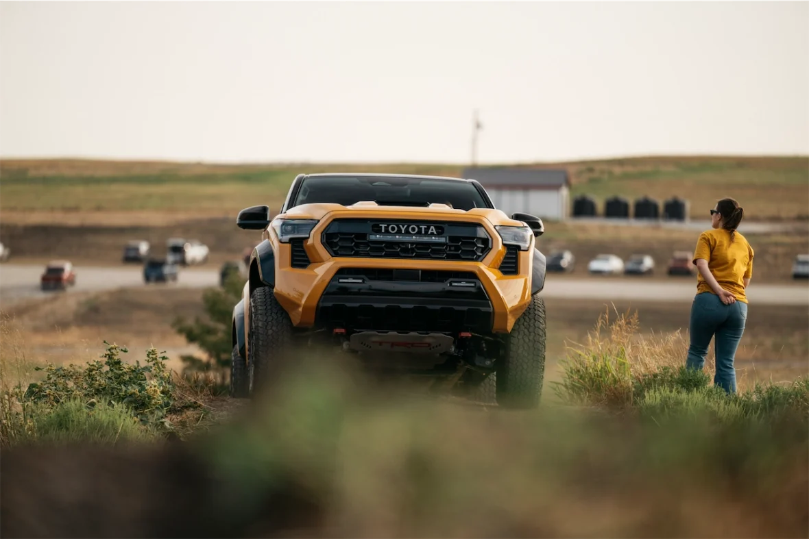 A yellow Toyota truck parked on a dirt path, with a woman in a yellow shirt standing nearby, looking towards the vehicle. The background features grassy hills and a parking area with other cars in the distance.