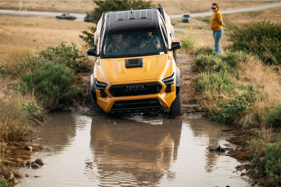 A bright yellow Toyota truck driving through a shallow muddy creek, with a woman standing nearby watching the vehicle.