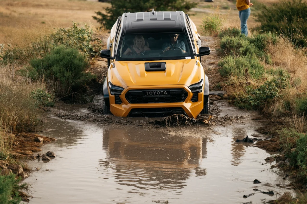 A yellow Toyota truck driving through a muddy water crossing in a natural landscape, with grass and bushes surrounding the area.