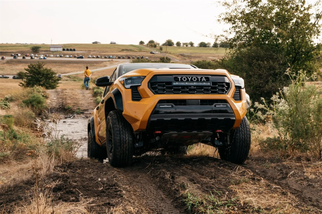 A yellow Toyota truck driving through an off-road path with dry grass and trees, while a person walks in the background.